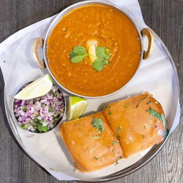 Pav Bhaji, a vibrant orange Indian curry, with butter, cilantro, chopped onions, and lime.