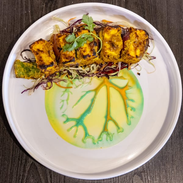 Grilled paneer tikka with cilantro, shredded cabbage, and artistic green and yellow sauces.