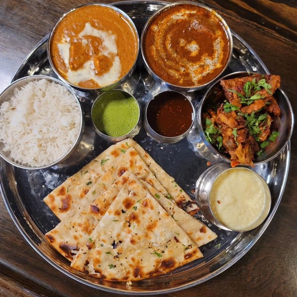 A Tap N Tikka Indian thali with curries, rice, naan, pakora, and sauces on a metal tray.