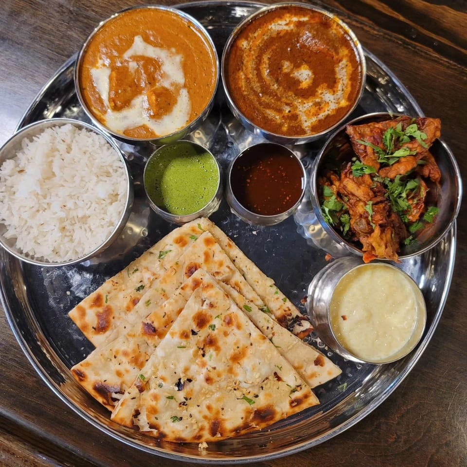 A Tap N Tikka Indian thali with curries, rice, naan, pakora, and sauces on a metal tray.